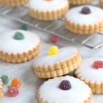 Empire Biscuits iced cookies, filled with jam and with scalloped edges topped with different coloured jelly tots, are arranged on a marble surface and cooling rack. Additional sweets are scattered around the biscuits.