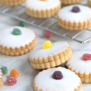 Empire Biscuits iced cookies, filled with jam and with scalloped edges topped with different coloured jelly tots, are arranged on a marble surface and cooling rack. Additional sweets are scattered around the biscuits.