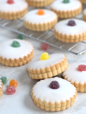 Empire Biscuits iced cookies, filled with jam and with scalloped edges topped with different coloured jelly tots, are arranged on a marble surface and cooling rack. Additional sweets are scattered around the biscuits.