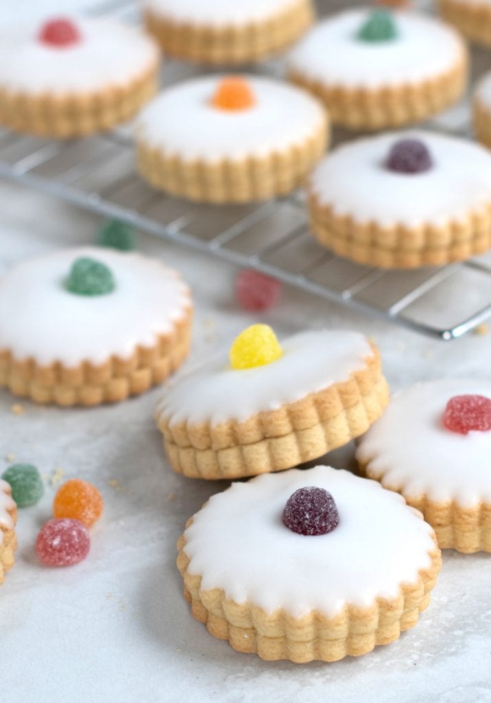 Empire Biscuits iced cookies, filled with jam and with scalloped edges topped with different coloured jelly tots, are arranged on a marble surface and cooling rack. Additional sweets are scattered around the biscuits.
