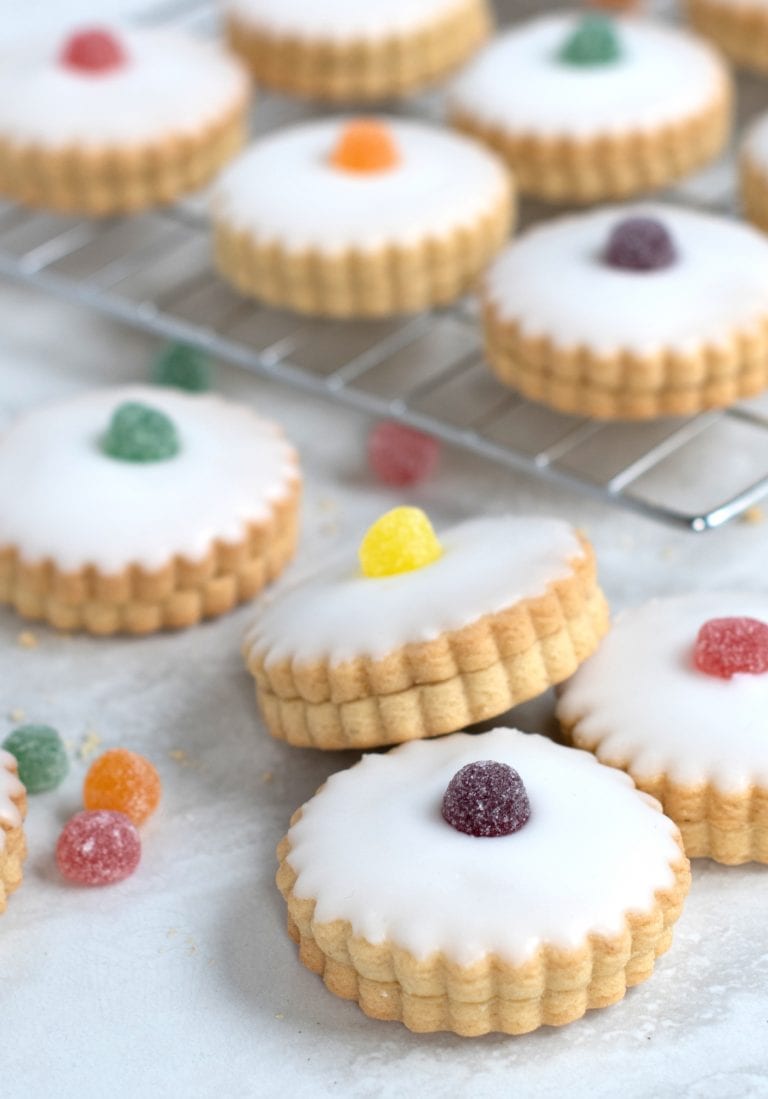 Empire Biscuits iced cookies, filled with jam and with scalloped edges topped with different coloured jelly tots, are arranged on a marble surface and cooling rack. Additional sweets are scattered around the biscuits.