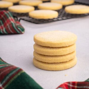 Stack of round Scottish Shortbread biscuits with more biscuits cooling on a wire rack behind.