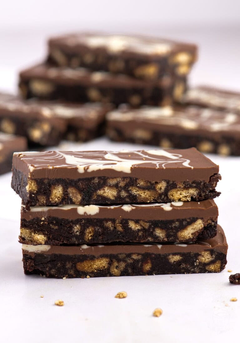 Stack of Scottish chocolate tiffin slices in focus, with a swirled chocolate topping and scattered biscuit pieces in the background.