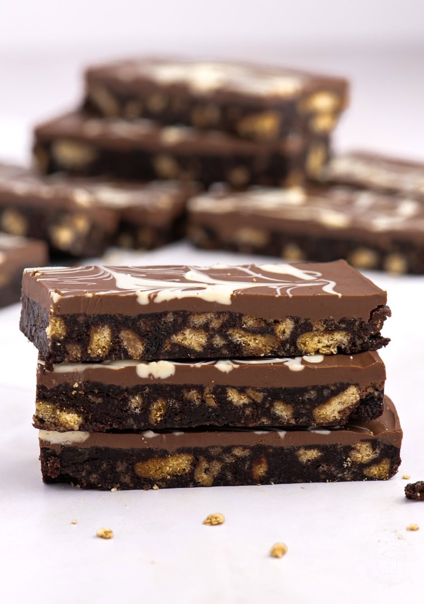 Stack of Scottish chocolate tiffin slices in focus, with a swirled chocolate topping and scattered biscuit pieces in the background.