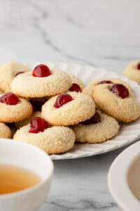 Coconut-coated Melting Moments topped with glacé cherries on a plate beside tea cups.