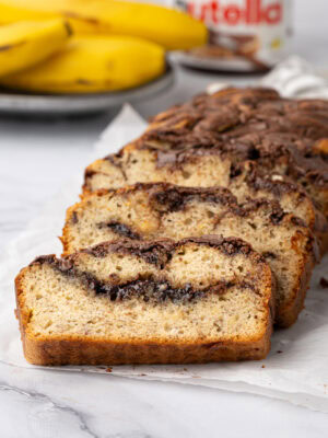 Banana Nutella loaf cake sliced on baking paper with bananas and a jar of Nutella behind.