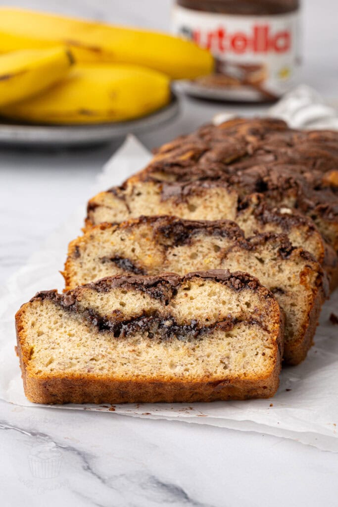 Banana Nutella loaf cake sliced on baking paper with bananas and a jar of Nutella behind.