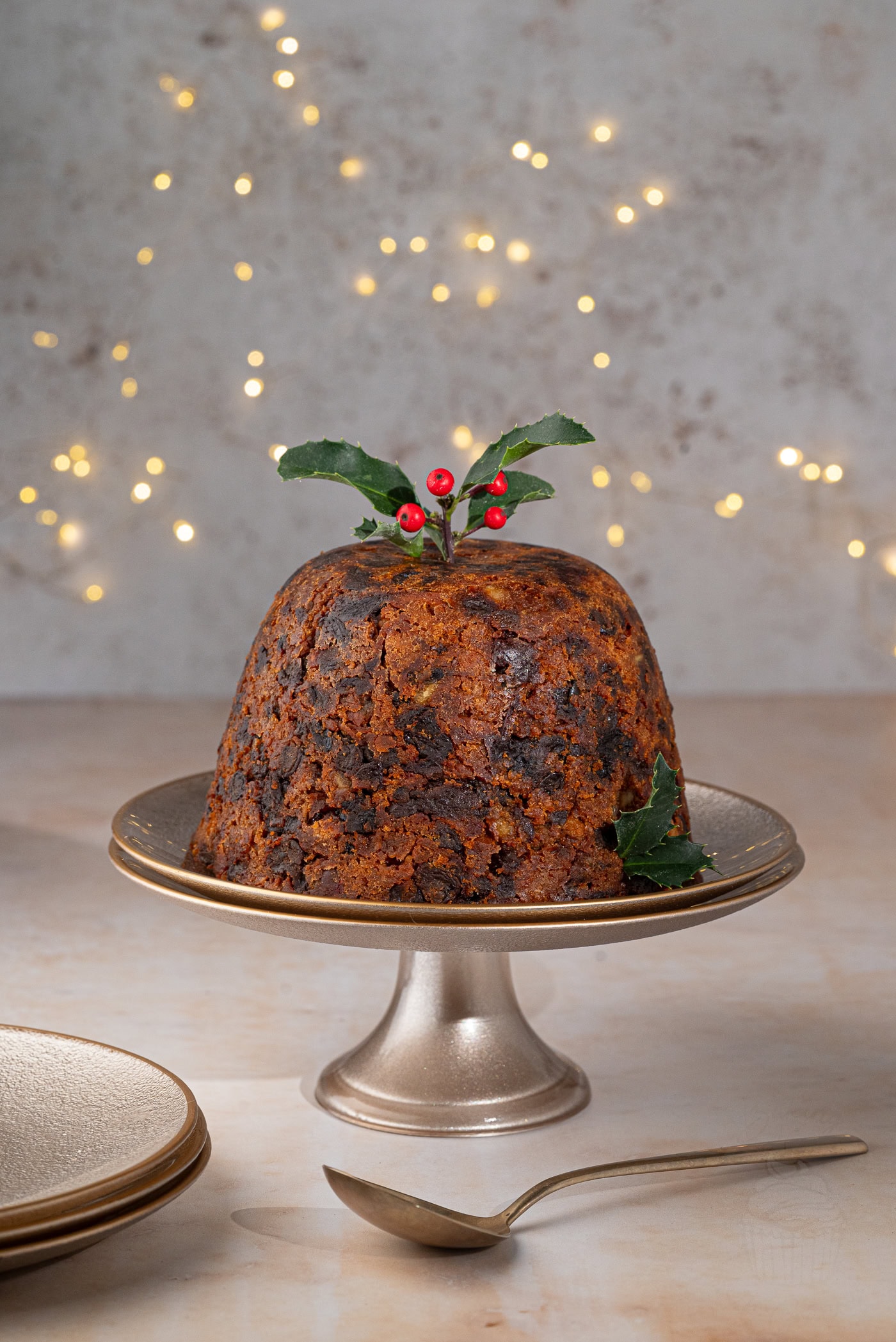A traditional British Christmas pudding topped with holly leaves and berries sits on a cake stand. The background features soft, glowing fairy lights for a warm holiday ambiance, capturing the spirit of a cherished recipe. A plate and spoon are placed nearby.