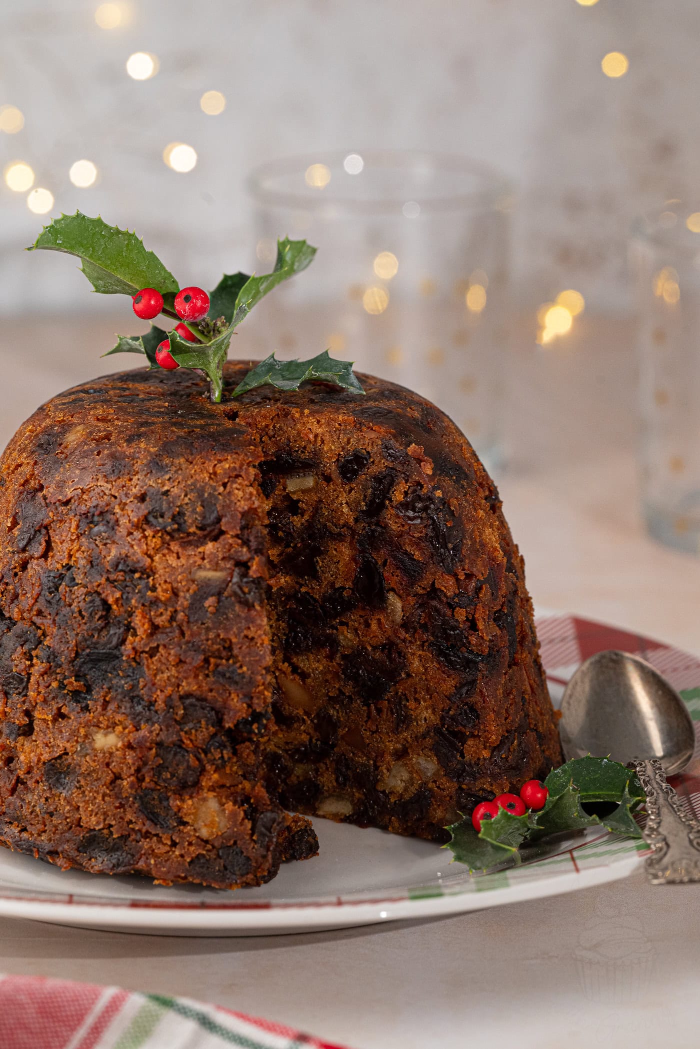 A festive Christmas pudding on a plate, garnished with holly leaves and red berries, captures the spirit of Stir-up Sunday. A slice has been cut, revealing its rich, fruit-filled texture. In the background, warm fairy lights create a cosy atmosphere. A spoon rests beside the pudding.