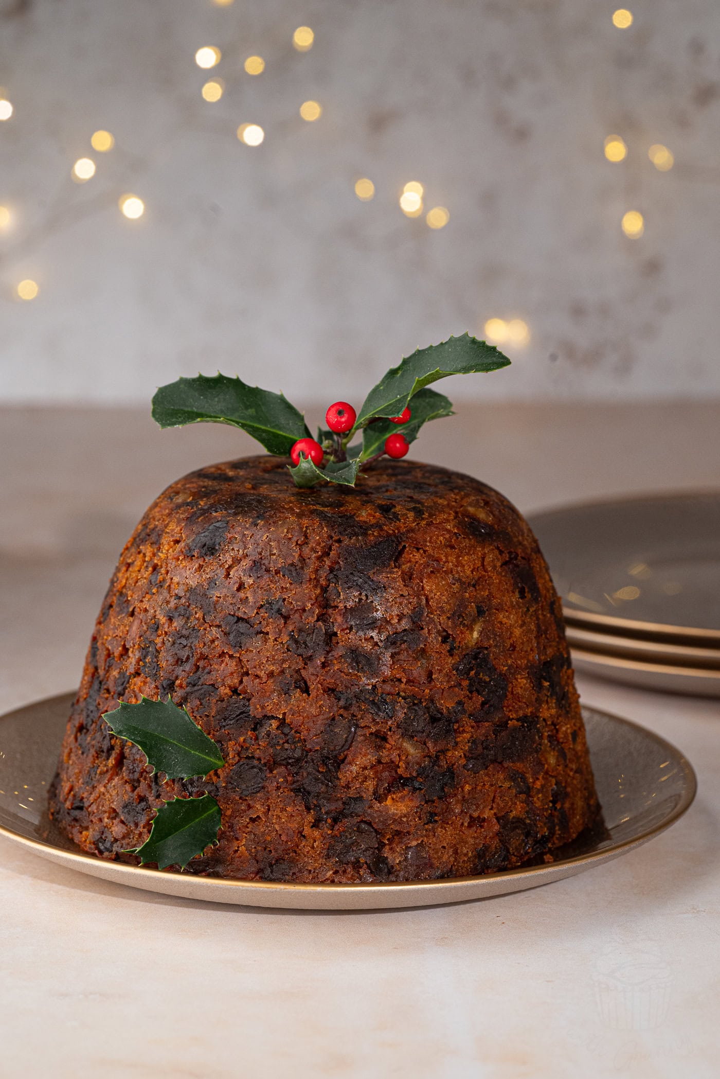 A traditional Christmas pudding, rich and dark, sits elegantly on a plate, adorned with holly leaves and red berries. The dessert is enveloped in the warm glow of soft, blurred festive lights, embodying the essence of holiday tradition.