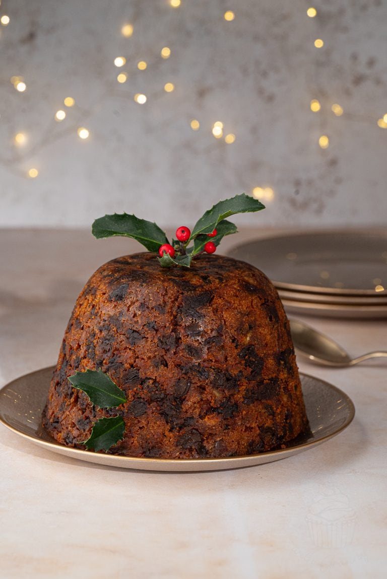 A traditional British Christmas pudding with a rich, dark texture sits on a plate. Its garnished with holly leaves and red berries. The background features soft, warm fairy lights and stacked plates.