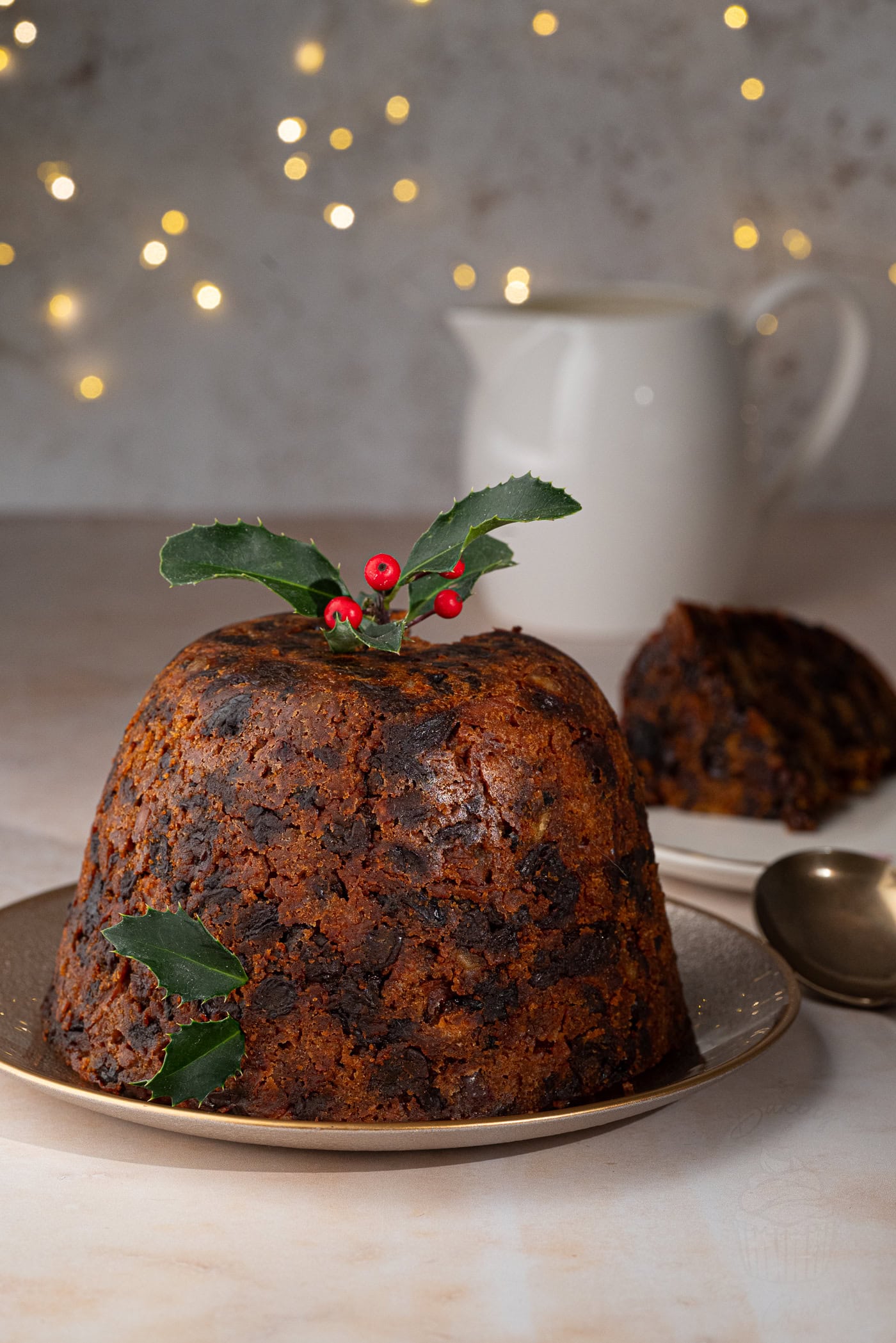 A homemade Christmas pudding decorated with holly sits on a plate. In the background, a piece of pudding is served on a dish, along with a cream pitcher. Fairy lights softly illuminate the scene, adding a festive touch.