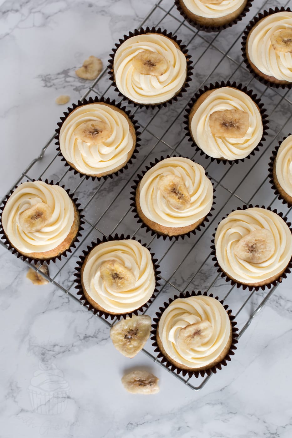 A dozen banana cupcakes, sat on a wire rack. Moist banana cupcakes, topped with vanilla buttercream and a banana chip.