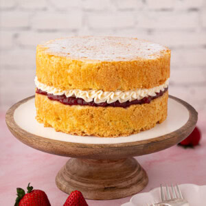 Full Victoria Sponge cake on a wooden stand, filled with strawberry jam and piped buttercream, surrounded by fresh strawberries.