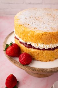 Close-up of a Victoria Sponge cake with visible layers of piped buttercream and jam, alongside whole strawberries on the cake stand.
