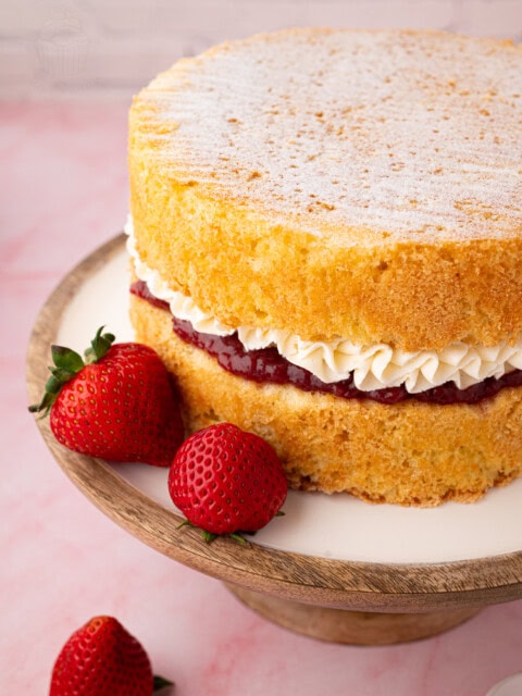 Close-up of a Victoria Sponge cake with visible layers of piped buttercream and jam, alongside whole strawberries on the cake stand.