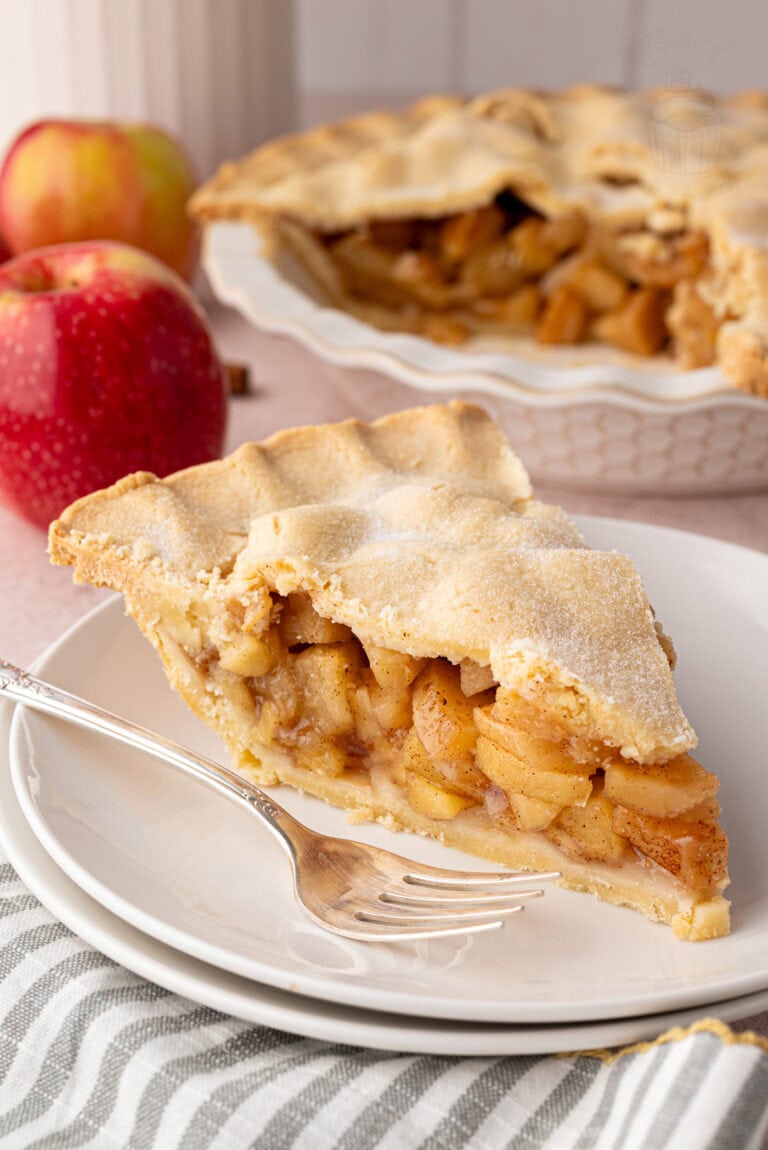 Slice of apple pie on a plate with the full pie in the background. A classic apple pie UK recipe made with sweet, spiced apples.