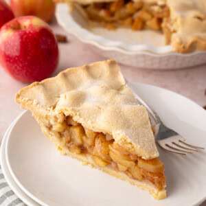 A slice of apple pie on a white plate, with fresh apples and the full pie visible in the background. A traditional apple pie UK recipe with shortcrust pastry.