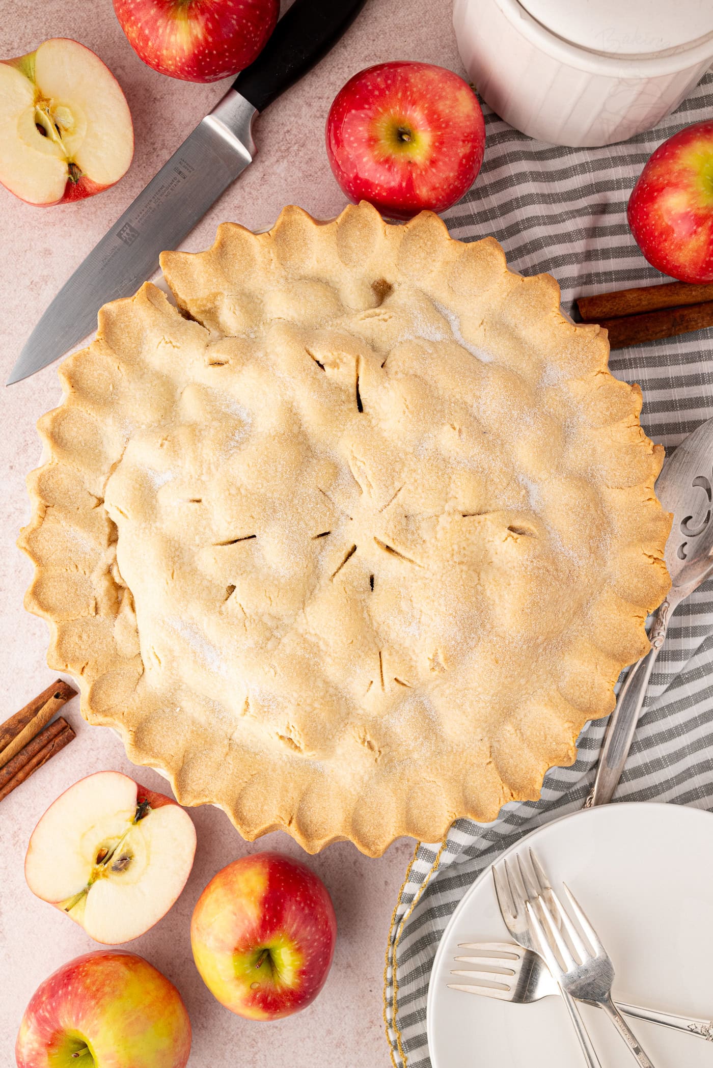 Top-down photo of a baked apple pie, styled with apples, cinnamon sticks, and cutlery. A seasonal apple pie UK recipe with classic flavours.