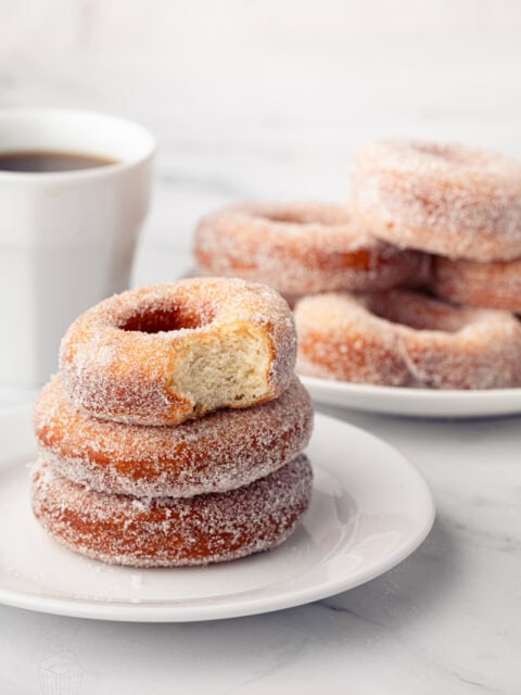 A stack of sugar-coated ring donuts on a small plate, with the top donut showing a bite taken out to reveal the fluffy texture inside.