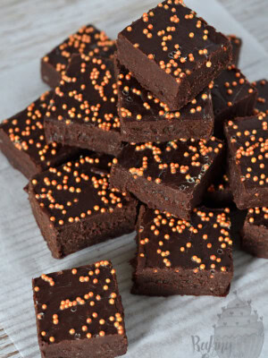 A stack of rich, sticky chocolate orange fudge topped with orange sprinkles, arranged neatly on parchment paper over a wooden surface. The vibrant sprinkles add a festive touch to the dark, fudgy goodness.