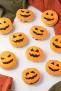 A group of orange-iced Pumpkin Empire Biscuits with black fondant pumpkin faces arranged on white surface.