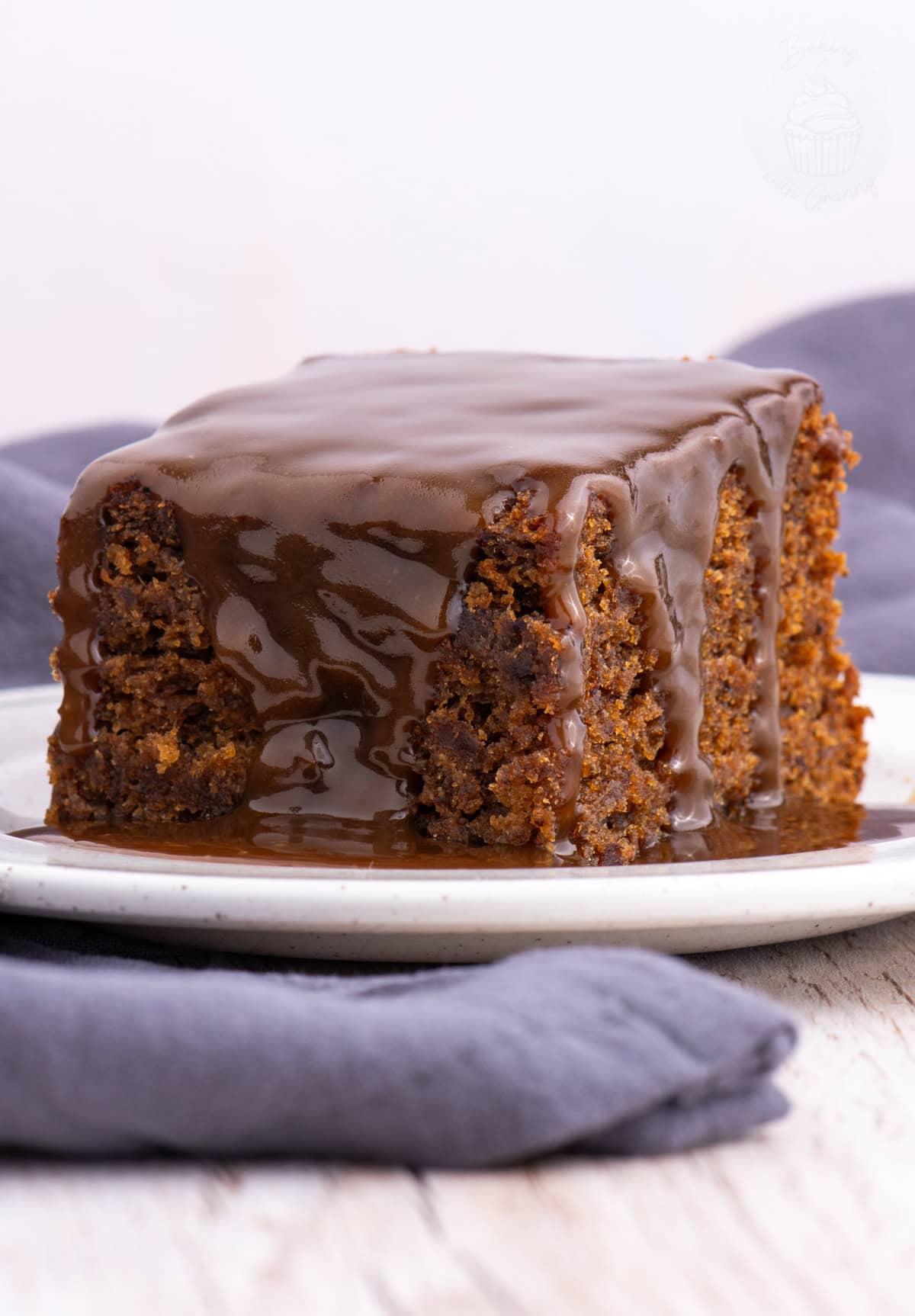 Slice of sticky toffee pudding on a plate, generously covered in glossy toffee sauce with sauce pooling underneath.