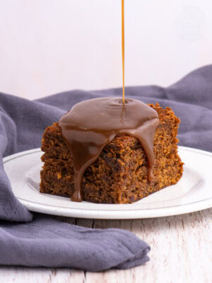 Warm sticky toffee pudding sponge with toffee sauce being poured over the top, dripping down the edges onto the plate.