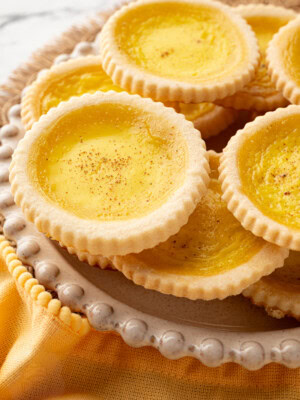 Custard tarts piled on a decorative plate with rustic woven placemat and mustard yellow napkin.
