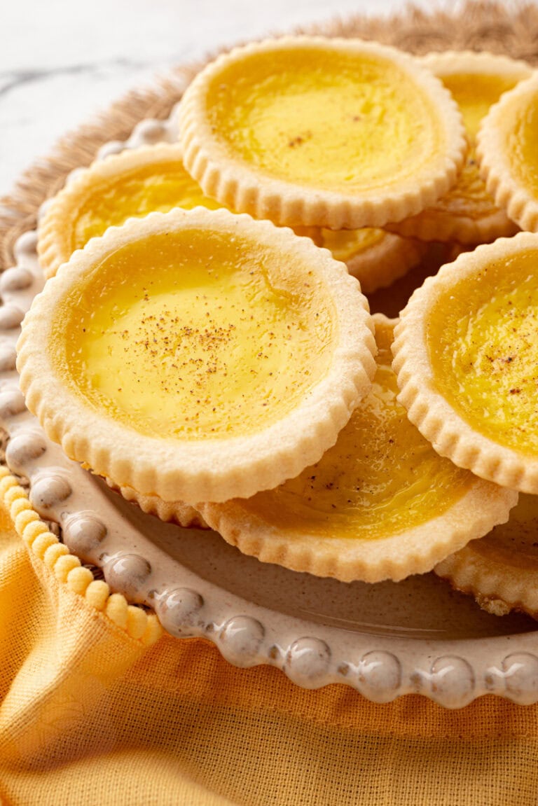Custard tarts piled on a decorative plate with rustic woven placemat and mustard yellow napkin.