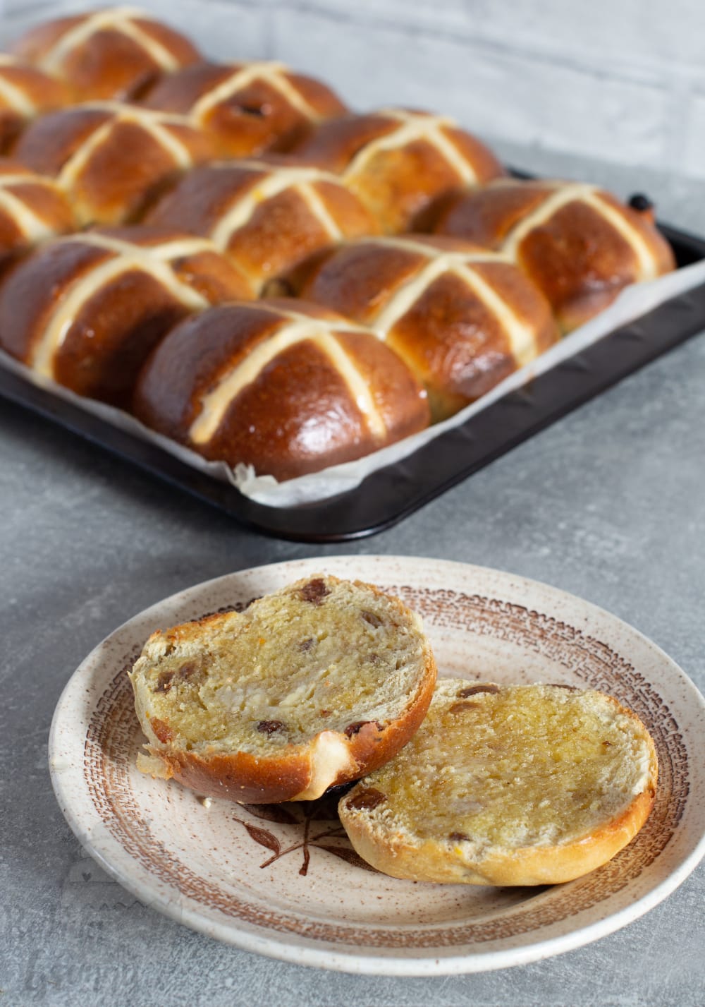 Homemade hot cross buns in baking tray with one sliced open and buttered showing soft fluffy texture.