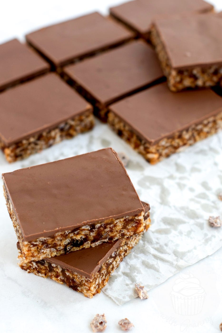 Two Krispy Date Slice squares on baking paper with chocolate topping and visible date and rice crispy texture.