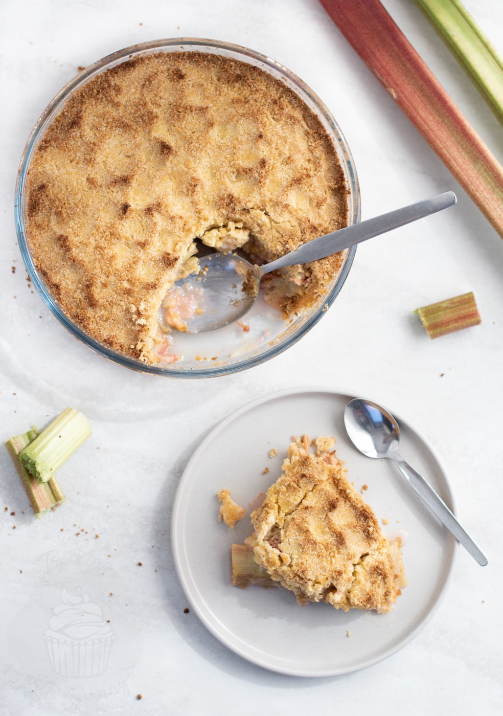 Overhead view of traditional rhubarb crumble in a baking dish with a serving on a plate beside it.