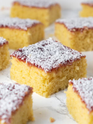 Slices of traditional jam and coconut school cake arranged on a white background, showing soft yellow sponge and coconut-covered jam topping.