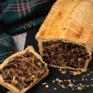 A Scottish black bun with a golden-brown crust rests on a black slate. One slice reveals a filling of dried fruit, nuts and raisins. A green and blue tartan cloth is in the background on a wooden surface.