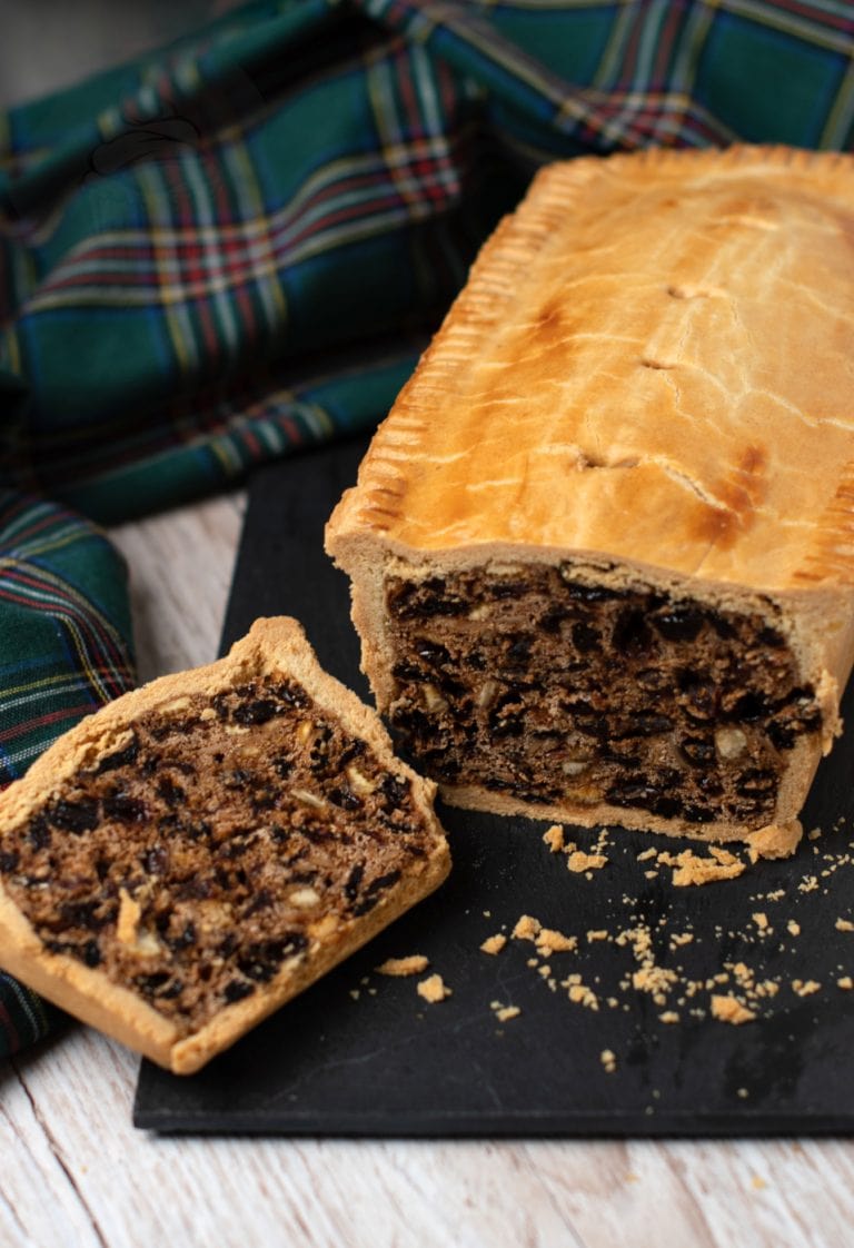 A Scottish black bun with a golden-brown crust rests on a black slate. One slice reveals a filling of dried fruit, nuts and raisins. A green and blue tartan cloth is in the background on a wooden surface.