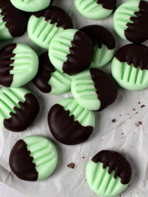A pile of round, green peppermint creams half-dipped in dark chocolate are scattered on crumpled white paper. These traditional peppermint creams have a grooved pattern on their tops. Chocolate crumbs speckle the paper
