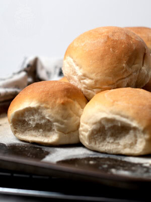 A close-up of freshly baked Scottish morning rolls on a baking tray. The rolls are golden brown with a slightly cracked, crusty surface. A white cloth is partially visible in the background, hinting at their homemade recipe origin.