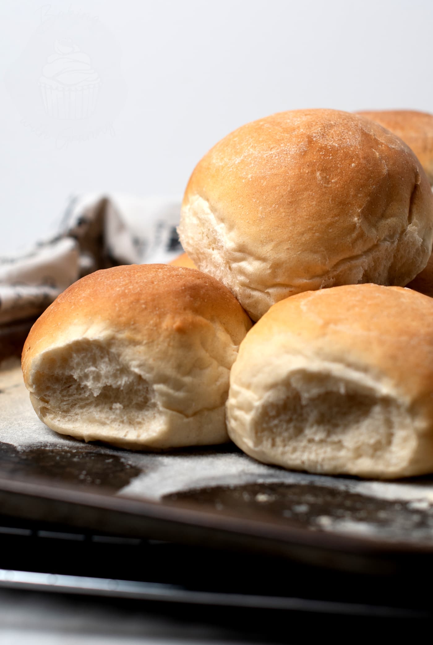 A close-up of freshly baked Scottish morning rolls on a baking tray. The rolls are golden brown with a slightly cracked, crusty surface. A white cloth is partially visible in the background, hinting at their homemade recipe origin.