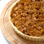 Cornflake tart upon a round wooden board, with a spoonful of strawberry jam behind.
