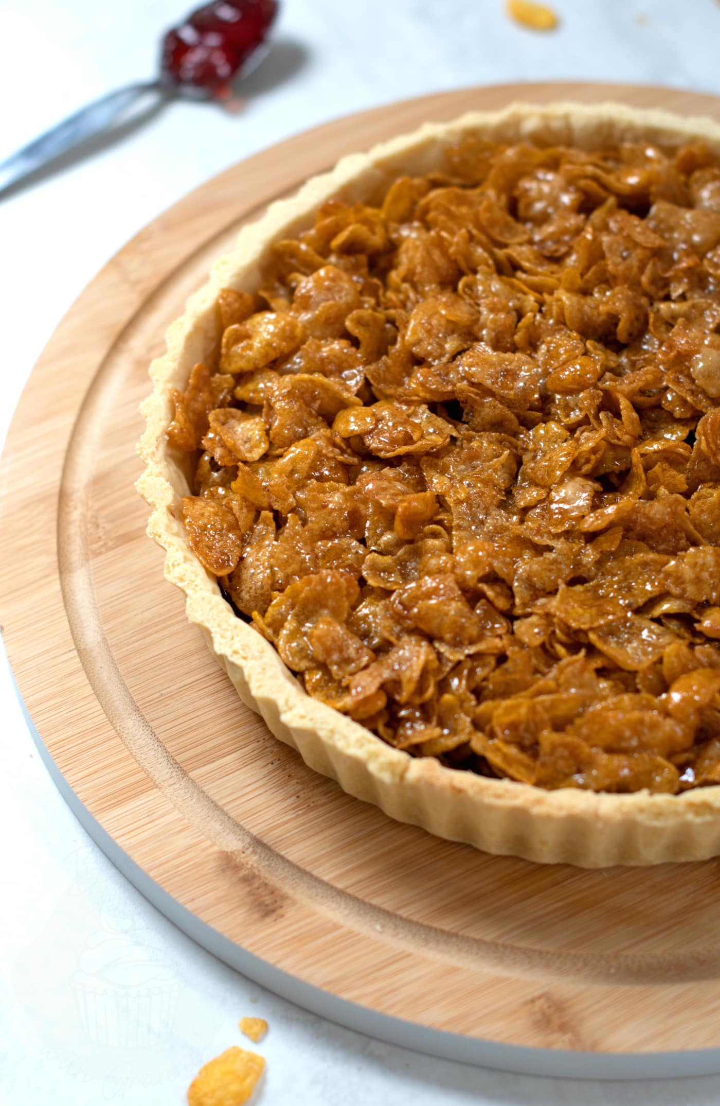 Cornflake tart upon a round wooden board, with a spoonful of strawberry jam behind.