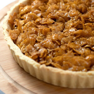 Close up of a golden cornflake tart, on a round wooden board, with a spoonful of jam in the background.