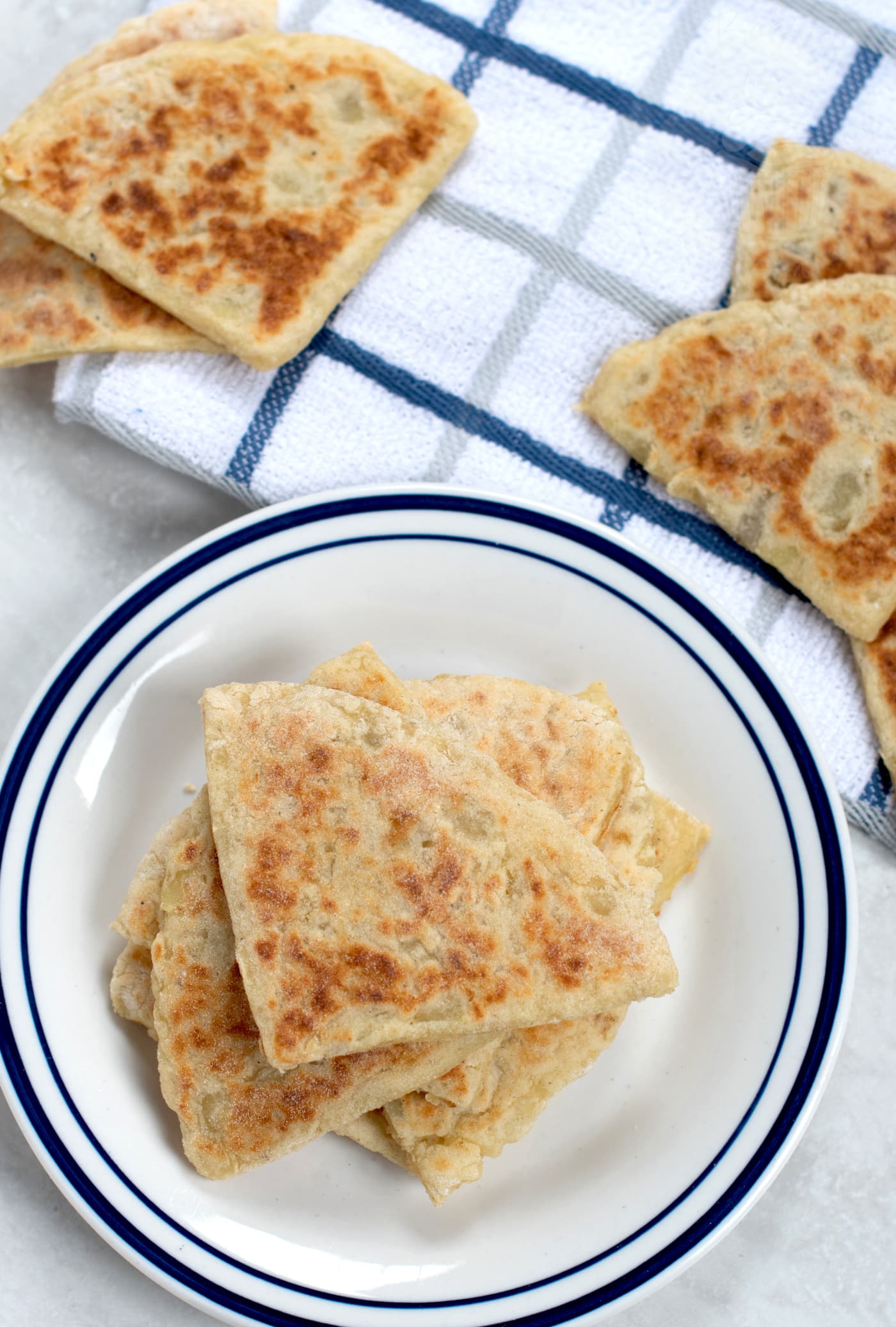 A plate of homemade tattie scones, with some more on the work surface beside. The plate is white with a blue rim, and there is also a white & blue tea towel on the work surface.