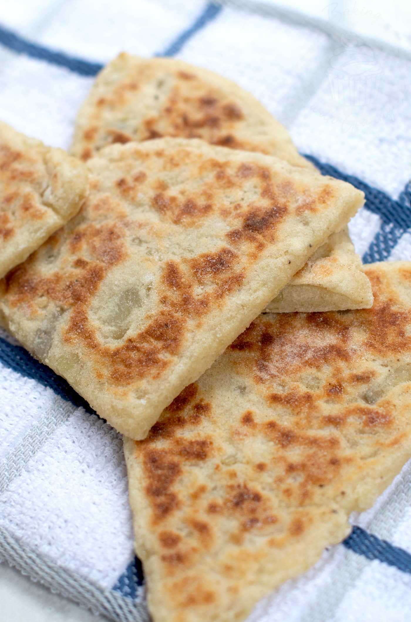Close view of a small pile of tattie scones. The golden browning on the surface of them can be seen. They are sat on top of a white & blue chequered tea towel.