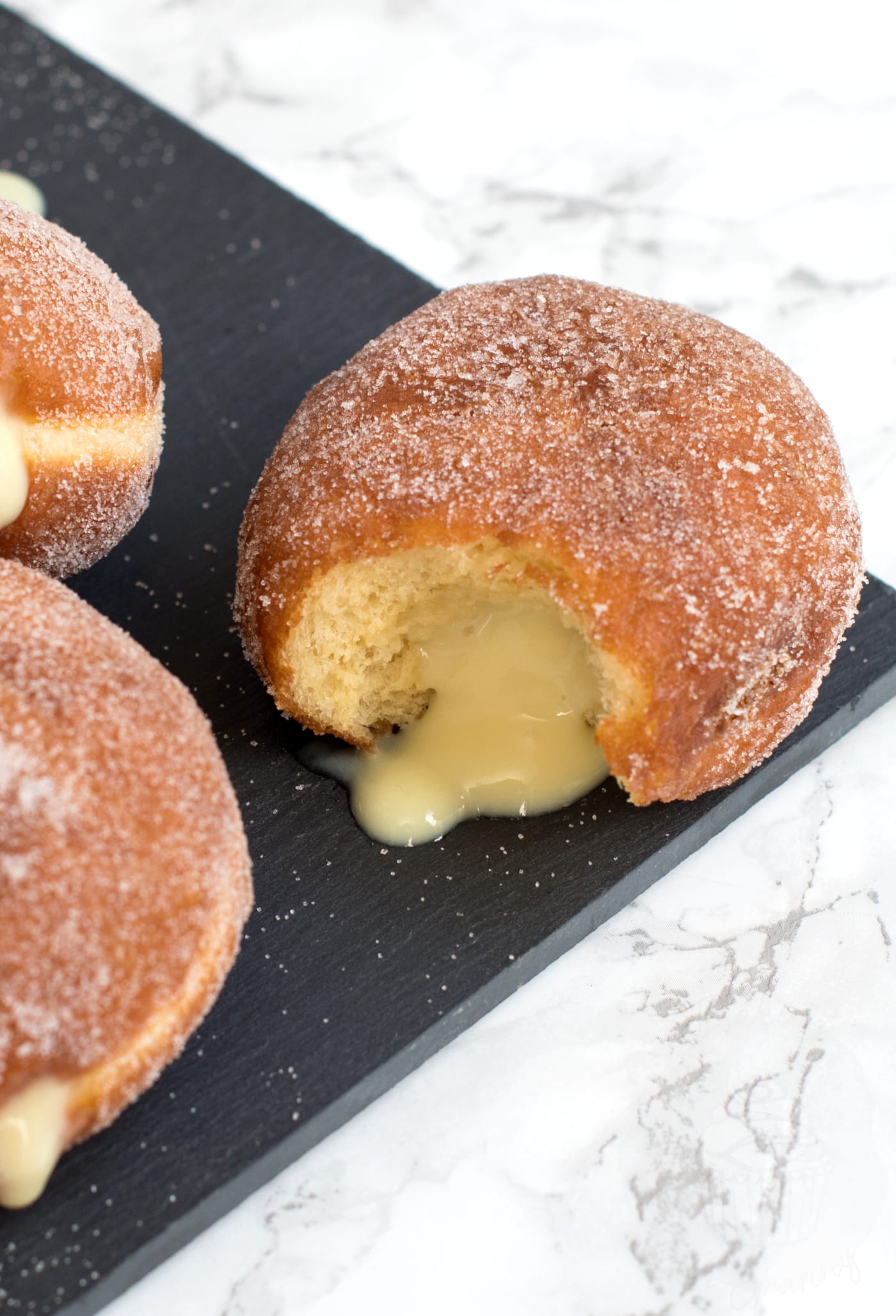 Custard doughnut with a bite out of it, with custard oozing from the middle. The doughnut is on a black slate, on top of white work surface.
