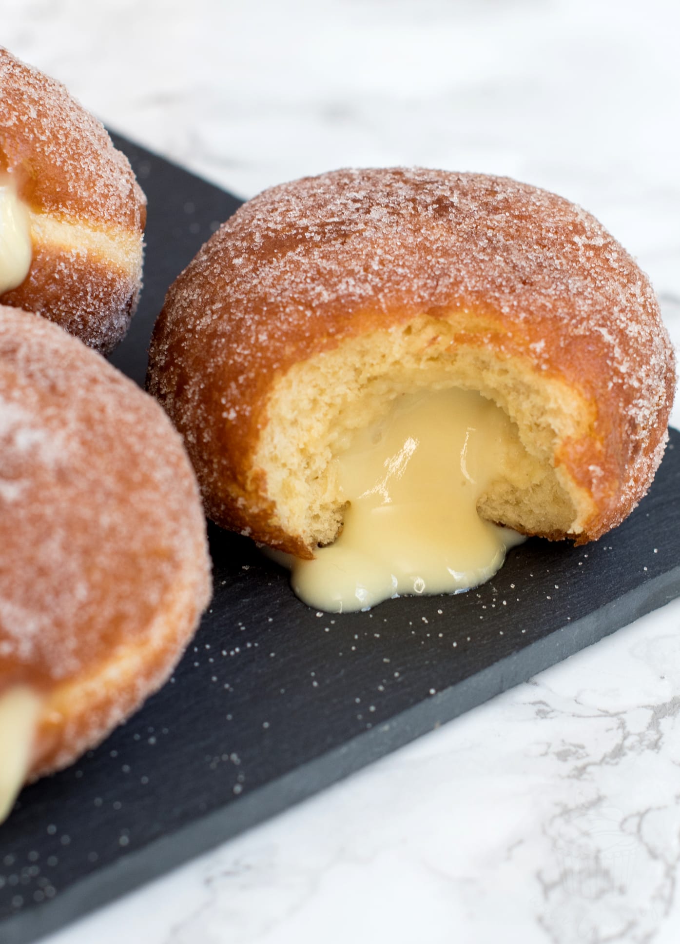 Custard donut with a bite out of it, with custard oozing from the middle. Whole donuts are seen beside.