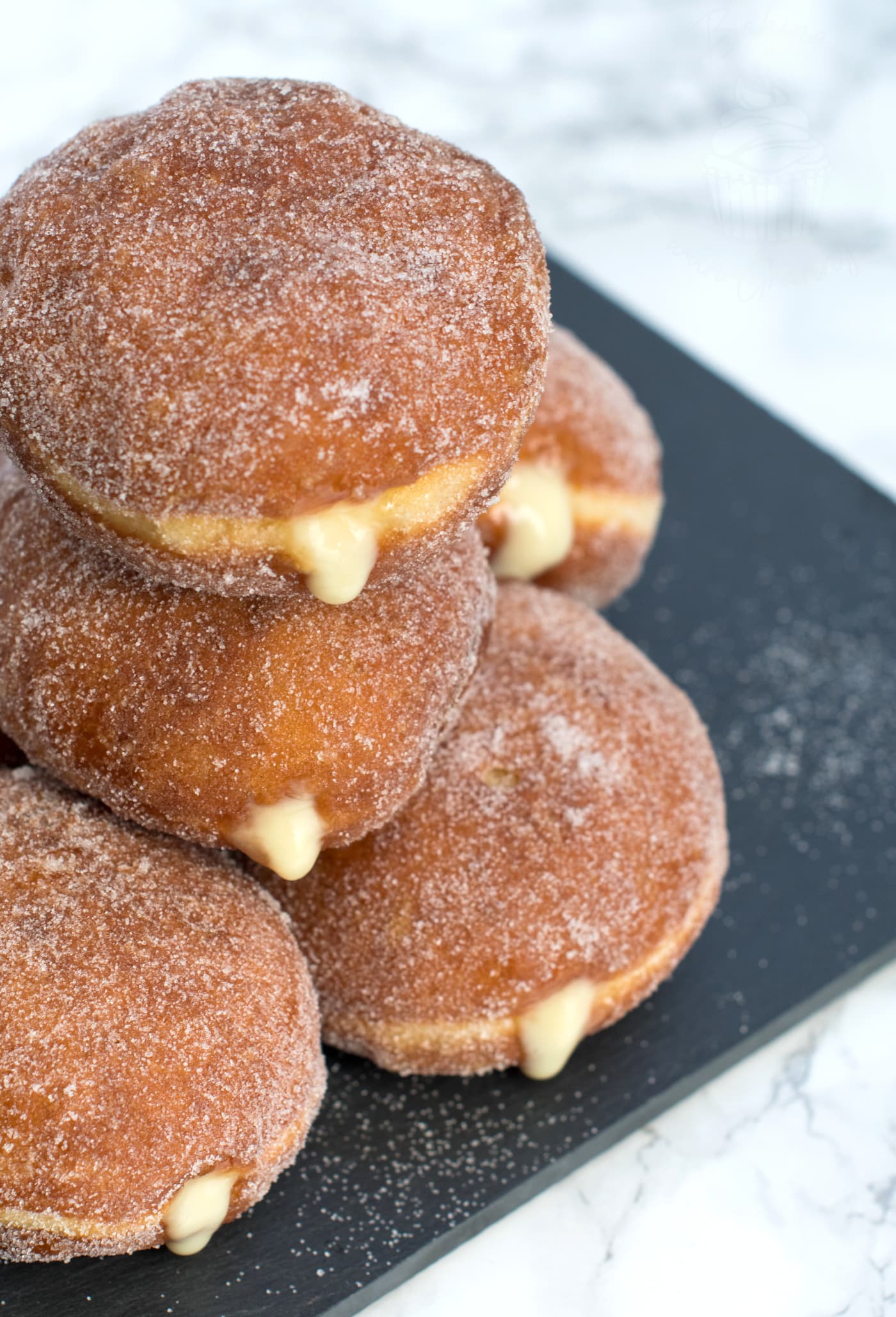 Stack of custard doughnuts, seen from the top. Custard is oozing from the doughnuts, which are golden in colour and covered in sugar.