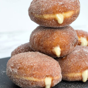 Stack of custard doughnuts on a black slate chopping board. The custard can be seen oozing from the sides of the sugar coated doughnuts.