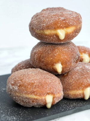 Stack of custard doughnuts on a black slate chopping board. The custard can be seen oozing from the sides of the sugar coated doughnuts.