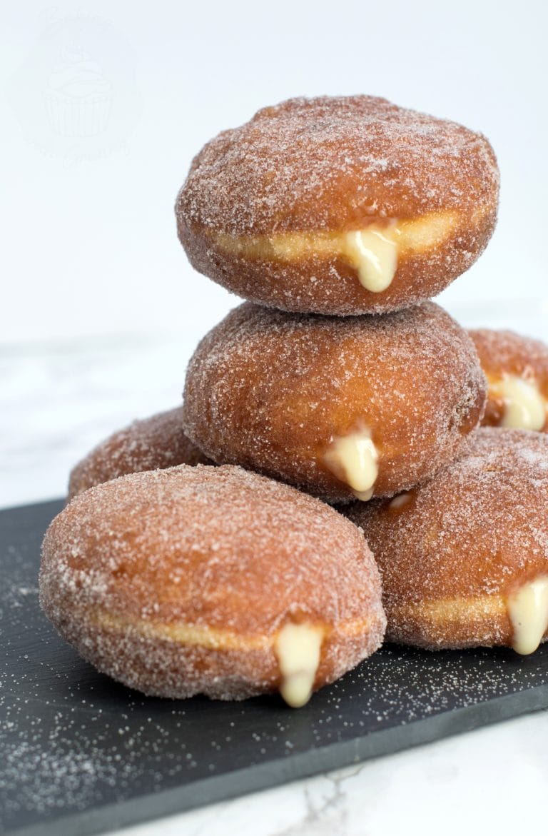 Stack of custard doughnuts on a black slate chopping board. The custard can be seen oozing from the sides of the sugar coated doughnuts.